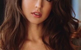 Portrait of a woman with long wavy brown hair and smoky makeup, looking at the camera, wearing an open green jacket.